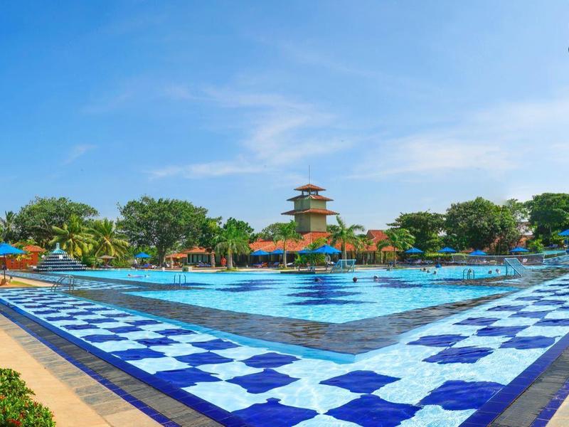 Large outdoor pool with blue and white pattern, surrounded by greenery and resort buildings under clear sky.
