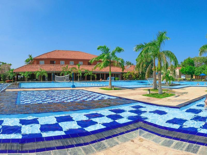 Large swimming pool with blue and white mosaic tiles, surrounded by palm trees and a hotel building.