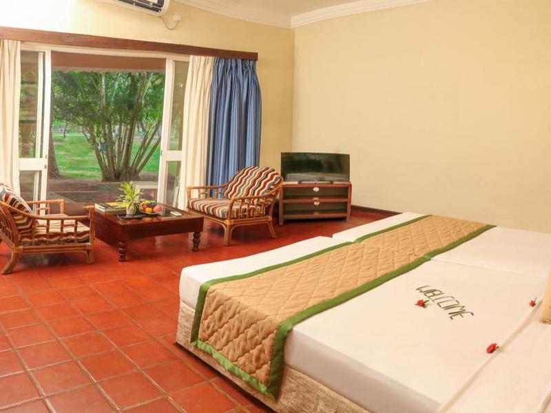 Bright hotel room with double bed, desk, chairs, and window overlooking green trees.