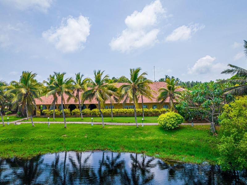 Hotel building behind palm trees and green lawn with pond in front under blue sky.