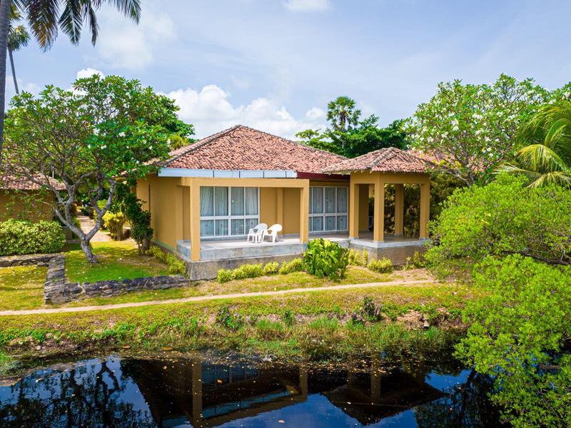 A yellow bungalow with a red tiled roof surrounded by green plants next to a small pond.