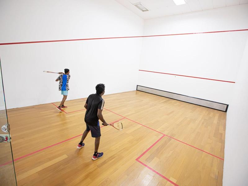 Two people playing squash in a bright, modern indoor squash court with wooden flooring.