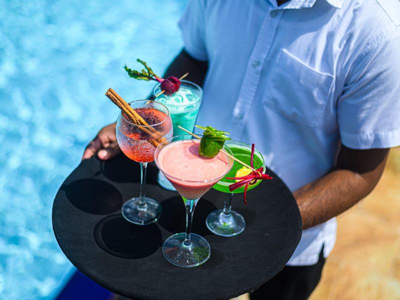 Waiter carrying tray with three colorful cocktails by a pool.