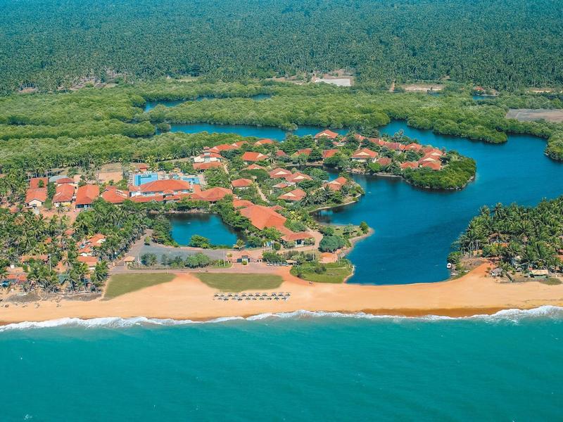 Aerial view of a resort with beach, lagoon, orange roofs, and dense forest in the background.
