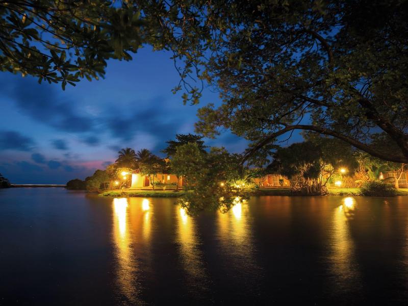 Night view of a lit hotel by the water with trees in the foreground.