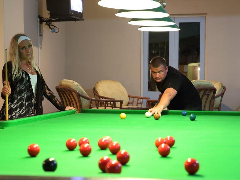 A couple plays billiards in a cozy room with chairs and a wall-mounted TV.