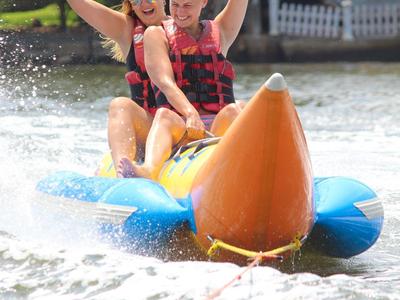 Zwei Personen sitzen auf einem gelben, orangefarbenen und blauen Bananenboot im Wasser und werfen die Arme in die Luft.