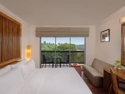 Helles Hotelzimmer mit großem Bett, Sofa, Holzdetails und Balkon mit Blick auf grüne Landschaft.