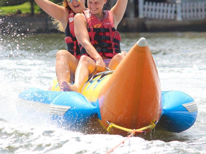 Zwei Personen im roten Schwimmwesten fahren auf einem blauen, orangen aufblasbaren Bananenboot.
