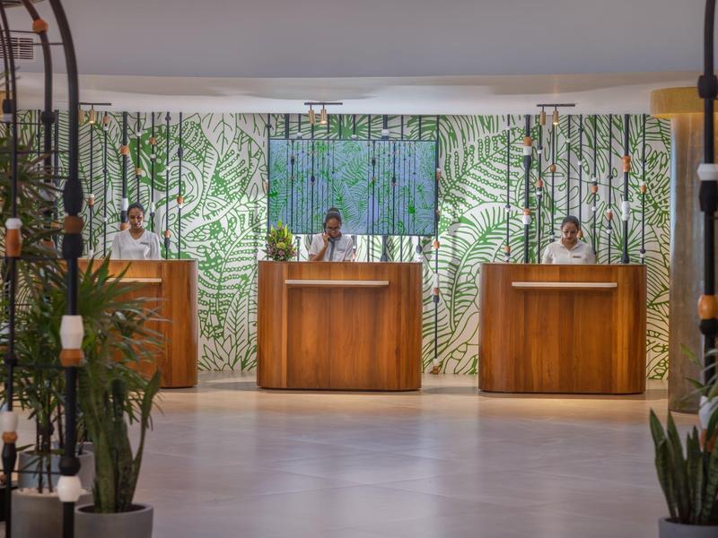 Moderne Hotelrezeption mit drei Holzpulten, grünem Tropenmuster-Hintergrund und Pflanzen im Vordergrund.