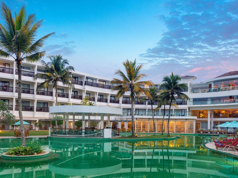 Moderner Hotelkomplex mit mehrstöckigem Gebäude, Palmen und großem, grünem Pool bei Abenddämmerung.