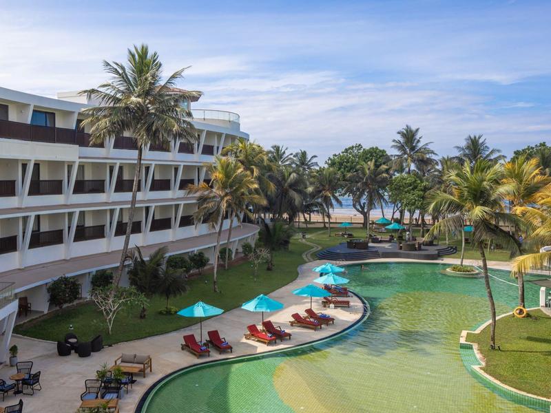 Luxushotel mit geschwungener Poolanlage, Liegestühlen, Palmen und Blick auf den blauen Himmel.