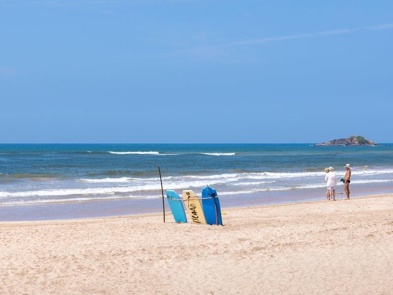 Strand mit hellem Sand, blauem Meer und wenigen Menschen im Hintergrund bei klarem Himmel.