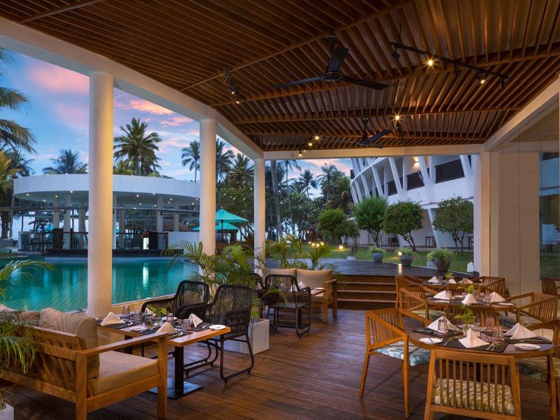Offenes Restaurant mit Holzboden, Rattanmöbeln neben einem beleuchteten Pool und Palmen bei Abendlicht.