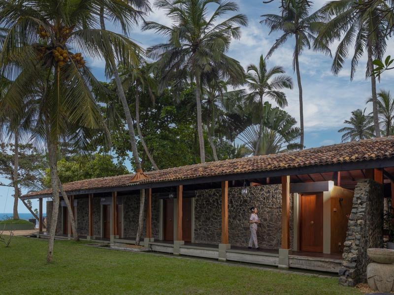 Langgestrecktes Gebäude mit Holzwänden und vielen Palmen davor unter bewölktem Himmel.