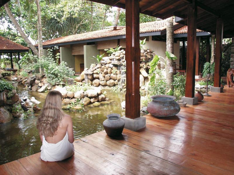 Frau sitzt auf Holzterrasse mit Blick auf Teich und Naturgarten mit Steinen und Pflanzen.
