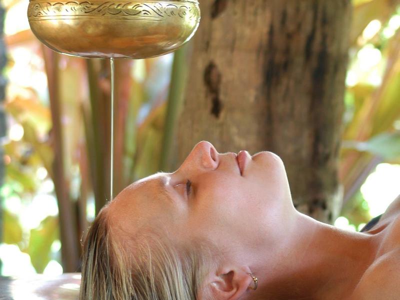 Frau legt mit geschlossenen Augen auf Holzbank, im Hintergrund Baum und sonnige Natur.