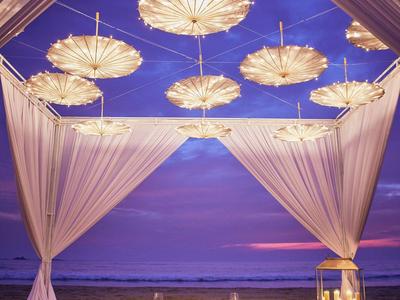 Romantische Strandlounge mit Kissen, Laternen und beleuchteten Sonnenschirmen bei Sonnenuntergang.