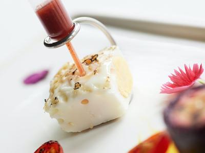 Ein Häppchen mit weißer Glasur und getrockneten Kräutern, versehen mit einer Pipette für eine rote Sauce.