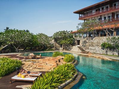 Hotelpool mit Sonnenliegen und umgeben von üppigem Grün unter klarem Himmel.