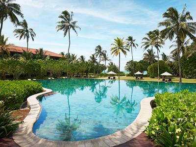 Großer Pool mit klarem Wasser, umgeben von Palmen und Sonnenschirmen in tropischer Hotelanlage.