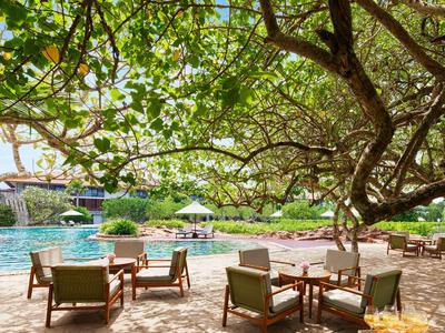 Sonnige Poollandschaft mit Liegestühlen und großen Bäumen für Schatten in einem Hotelresort.