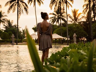 Una mujer está junto a una piscina con palmeras y sombrillas al atardecer.