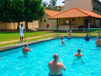 Menschen spielen Volleyball im Außenpool neben einem Hotelgebäude mit roten Dächern.
