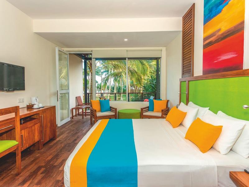 Jasny pokój hotelowy z podwójnym łóżkiem, kolorowymi poduszkami i tarasem z tropikalnym widokiem.