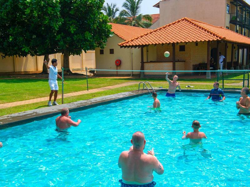 Ludzie ćwiczą aerobik w wodzie na zewnętrznym basenie przed budynkiem z czerwonym dachem.