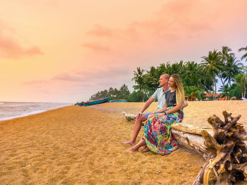 Para siedzi na kłodzie drewna na plaży, z zachodem słońca i palmami w tle.