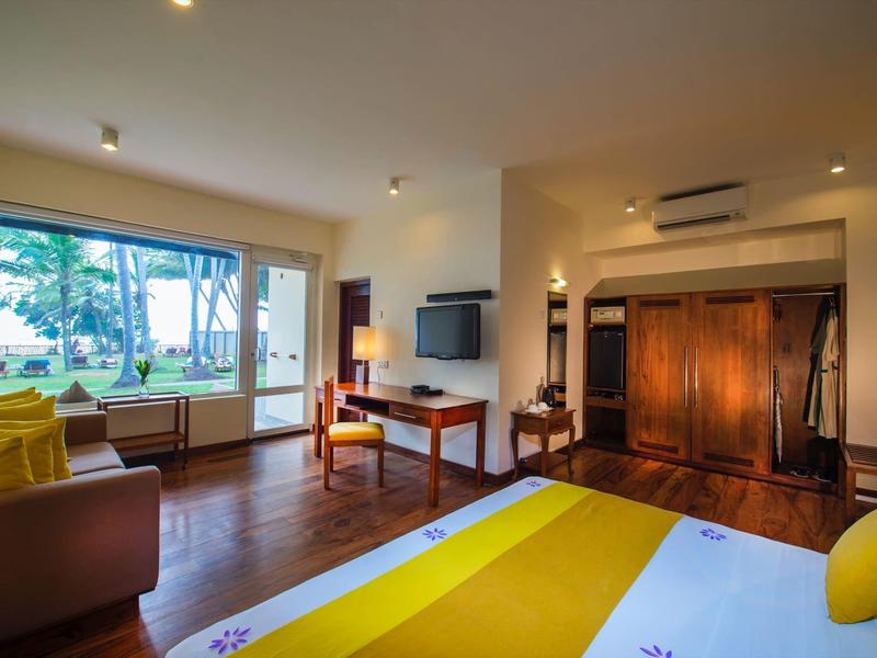 Jasny pokój hotelowy z żółtym łóżkiem, stołem, krzesłami i dużym oknem z widokiem na drzewa.