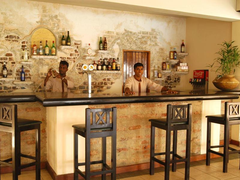 Bar z czterema wysokimi stołkami i dwoma barmanami przed rustykalną ścianą z półkami pełnymi butelek.