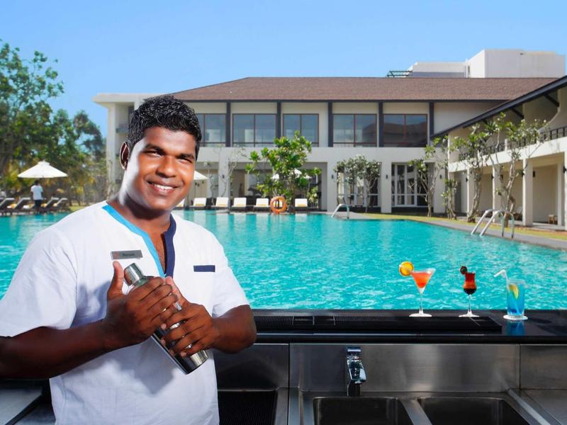 Barman souriant tenant une bouteille près de la piscine de l'hôtel avec bâtiment en arrière-plan.
