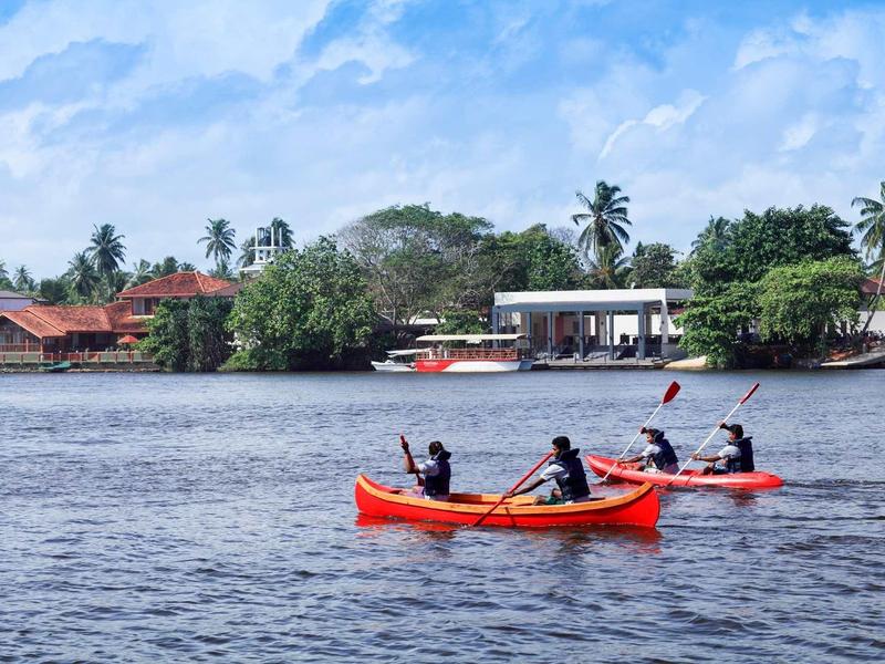Deux personnes pagaient en kayaks rouges sur une rivière avec des bâtiments et des palmiers en arrière-plan.