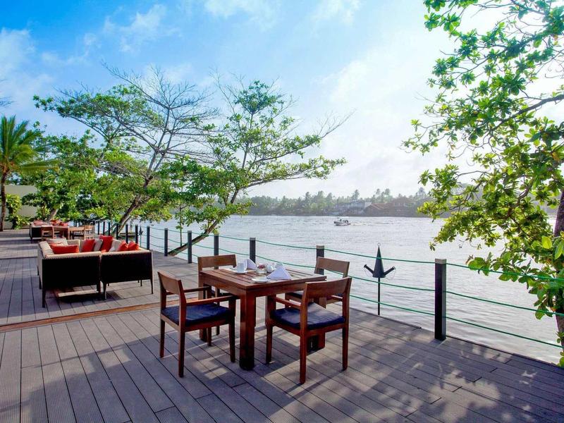 Terrasse avec tables et chaises au bord d'une rivière, entourée d'arbres avec vue sur la ville en arrière-plan.
