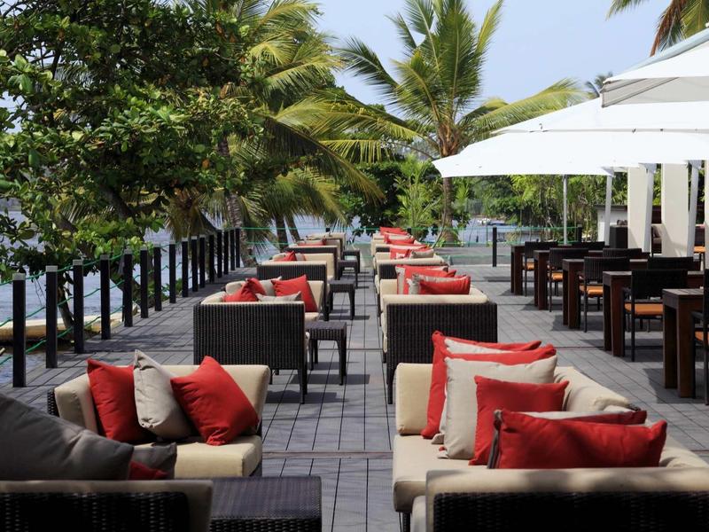 Salon en plein air avec canapés confortables, coussins rouges et parasols blancs au bord de l'eau.