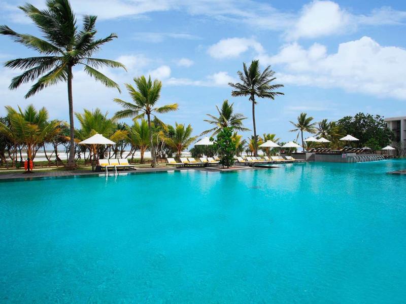 Grande piscine avec palmiers et parasols sous un ciel bleu dans la station hôtelière.