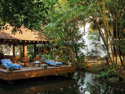 Overdekt houten terras met ligstoelen bij het water, omgeven door groene bomen en bamboe.