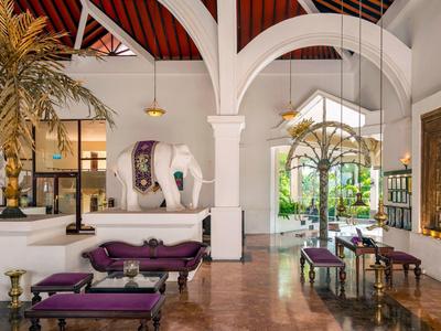 Lichte hotellobby met witte muren, hoge gewelfde plafonds, palmbomen en paarse zitmeubels.