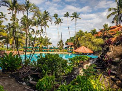 Tropischer Hotelpool mit Palmen, üppiger Vegetation und blauem Himmel.