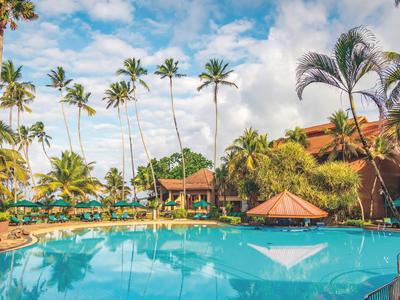 Großer Pool vor einem tropischen Hotel mit Palmen und blauem Himmel.