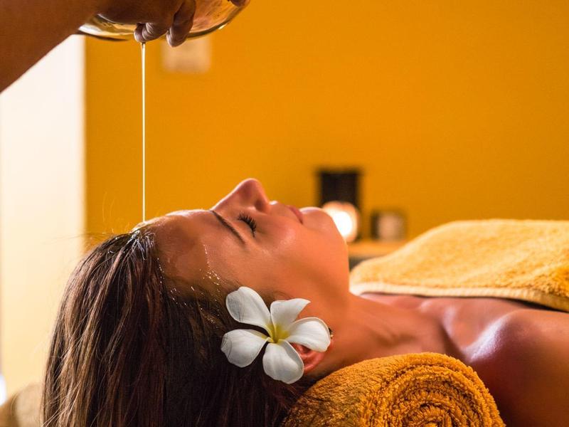 Vrouw ontspant tijdens Ayurvedische hoofdmassage met verse bloem in haar en warme gele handdoek.