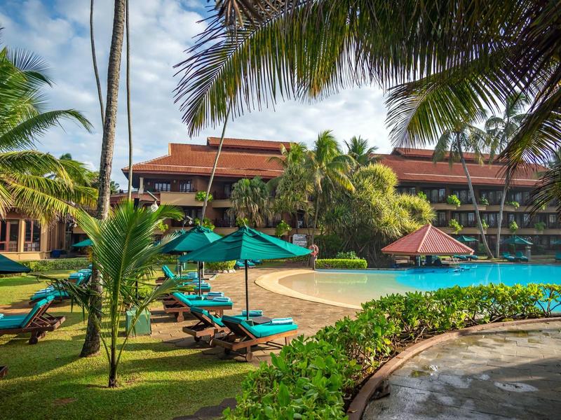 Hotelzwembad met ligstoelen, parasols en palmbomen onder een blauwe lucht.