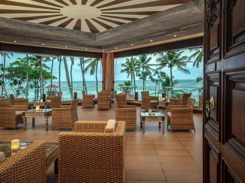 Open lounge met rotan meubels met uitzicht op palmbomen en oceaan bij zonsondergang.