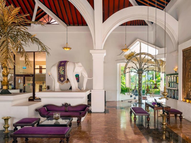 Lichte hotellobby met witte muren, hoge gewelfde plafonds, palmbomen en paarse zitmeubels.