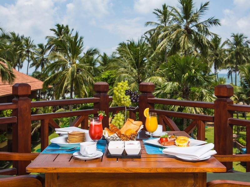 Tropische ontbijttafel met vers fruit en sappen op een terras met uitzicht op palmbomen.