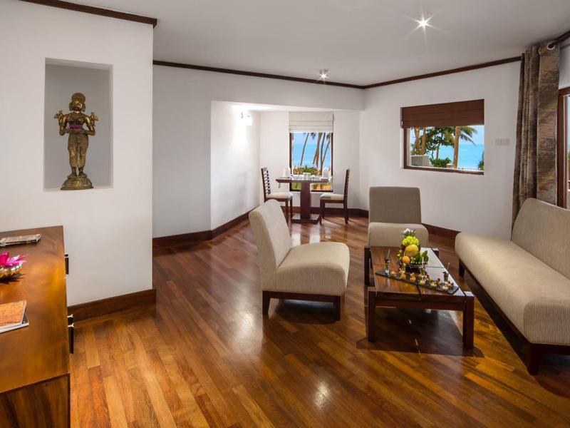 Woonkamer met houten vloer, bank, fauteuils en grote ramen met uitzicht op bomen.