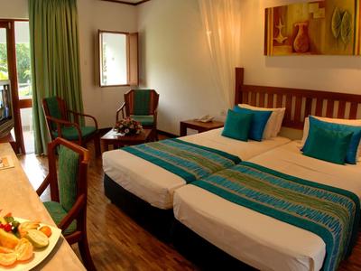 Světý hotelový pokoj se dvěma jednolůžky, modrými polštáři a přístupem na balkon s výhledem na zeleň.