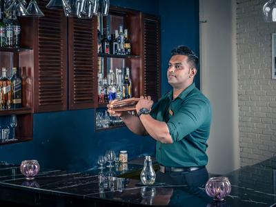 Barman připravující drink za elegantním barem z tmavého dřeva s osvětlením.